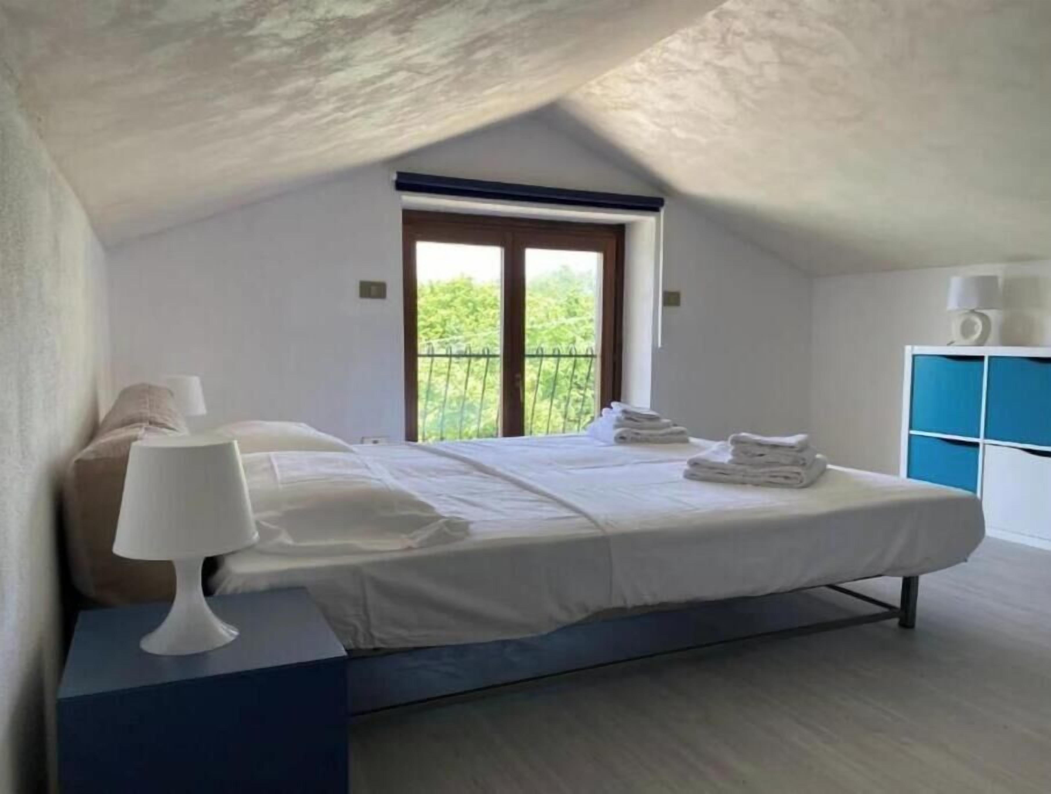 Un dormitorio con cama, lámpara de noche, ventana y vistas al verdor.