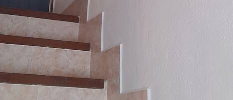 Un escalier aux marches beiges et aux murs blancs, menant à une porte sombre sur laquelle est fixée une applique lumineuse.