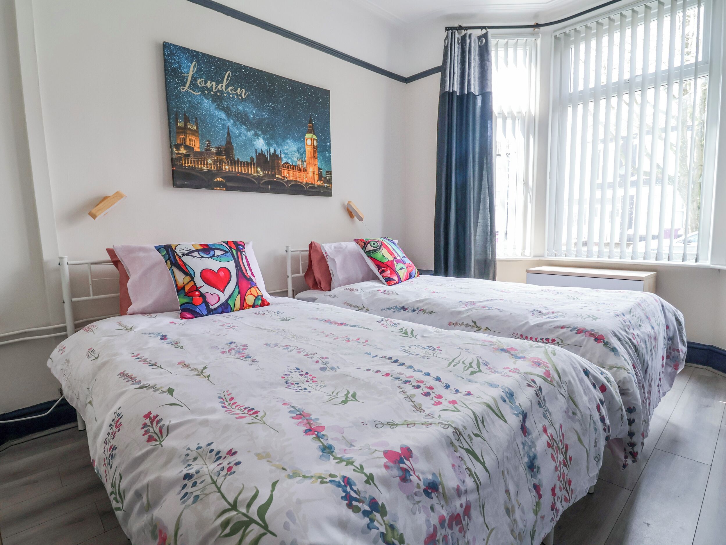 Una camera d'albergo con due letti, biancheria da letto floreale, un'opera d'arte con una veduta urbana e una finestra con tende.