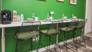 Un espace repas agrémenté d’un mur vert, de tableaux encadrés, d’une machine à café et de tabourets de bar.