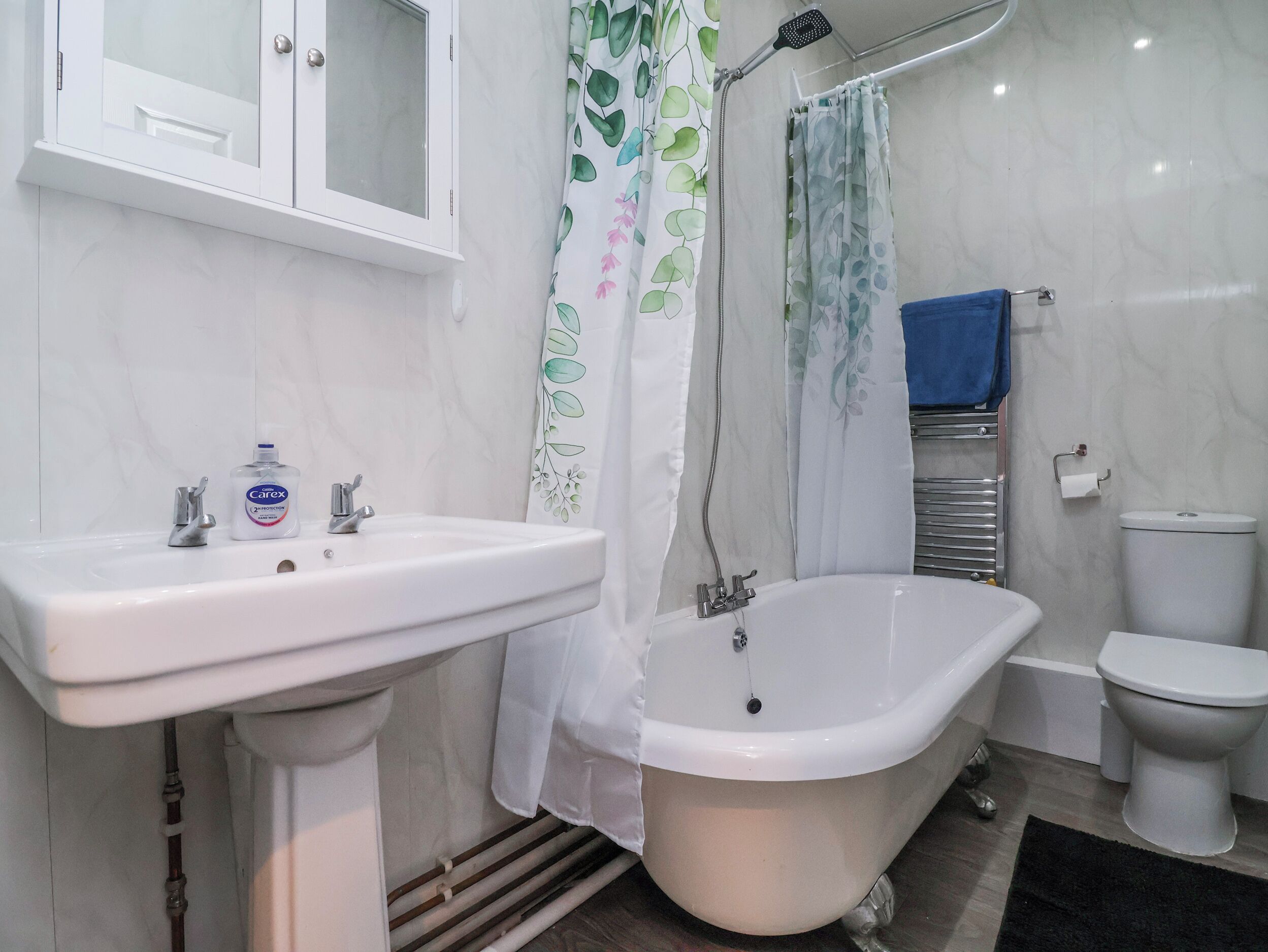 Une salle de bain avec un lavabo sur colonne, une baignoire, des toilettes et un rideau de douche orné d’un motif de feuilles.