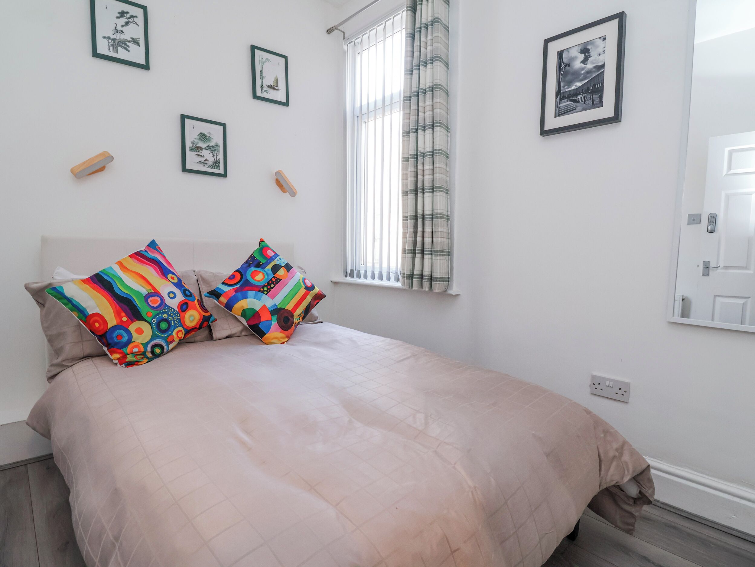 Un dormitorio con cama, almohadas, una ventana con cortinas y cuadros colgados en la pared.