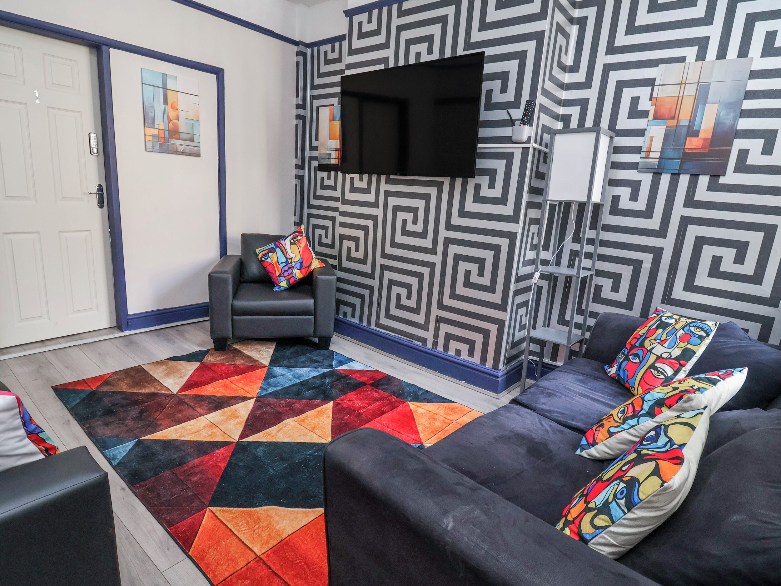 Una sala de estar con un sofá, un sillón, un tapete colorido y un televisor instalado en la pared.