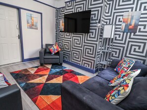 Una sala de estar con un sofá, un sillón, un tapete colorido y un televisor instalado en la pared.