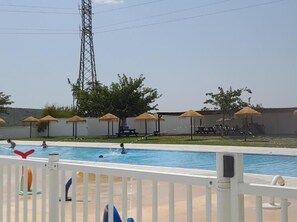 Una piscina con valla blanca, sombrillas y una zona de picnic al fondo.