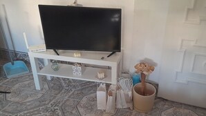 A room with a flat-screen TV on a white stand, a small white table with decorative items, a blue chair, and a patterned floor. - Roof Terrace - Pet - Parking - Pool- Sleeps 2 (Rute)