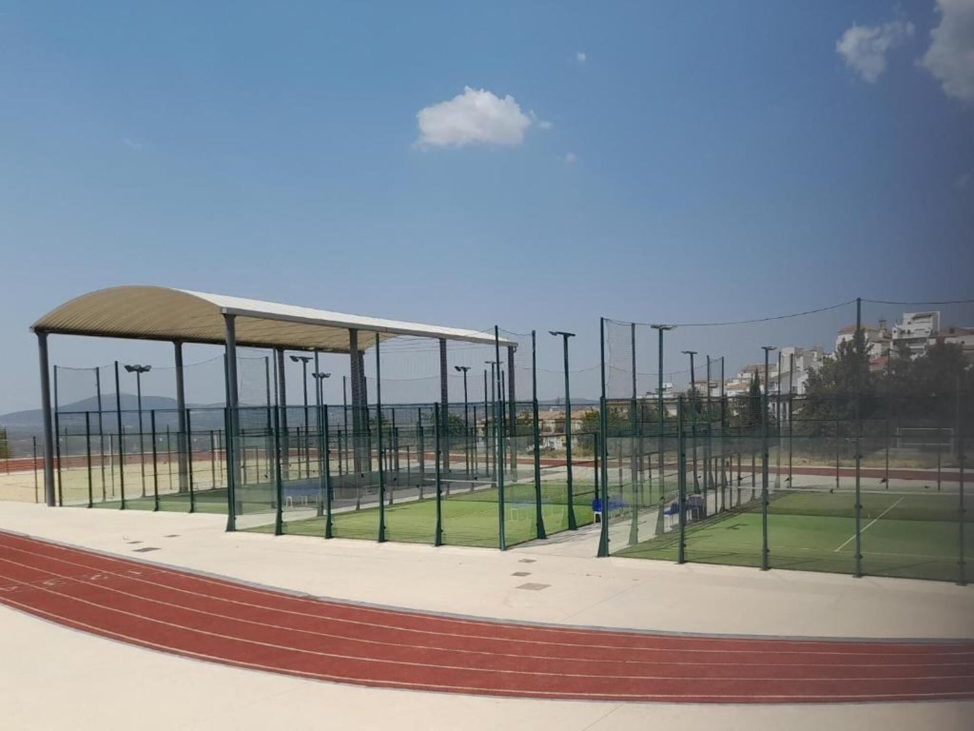 A sports facility with a running track, a covered seating area, and a netted tennis court.
