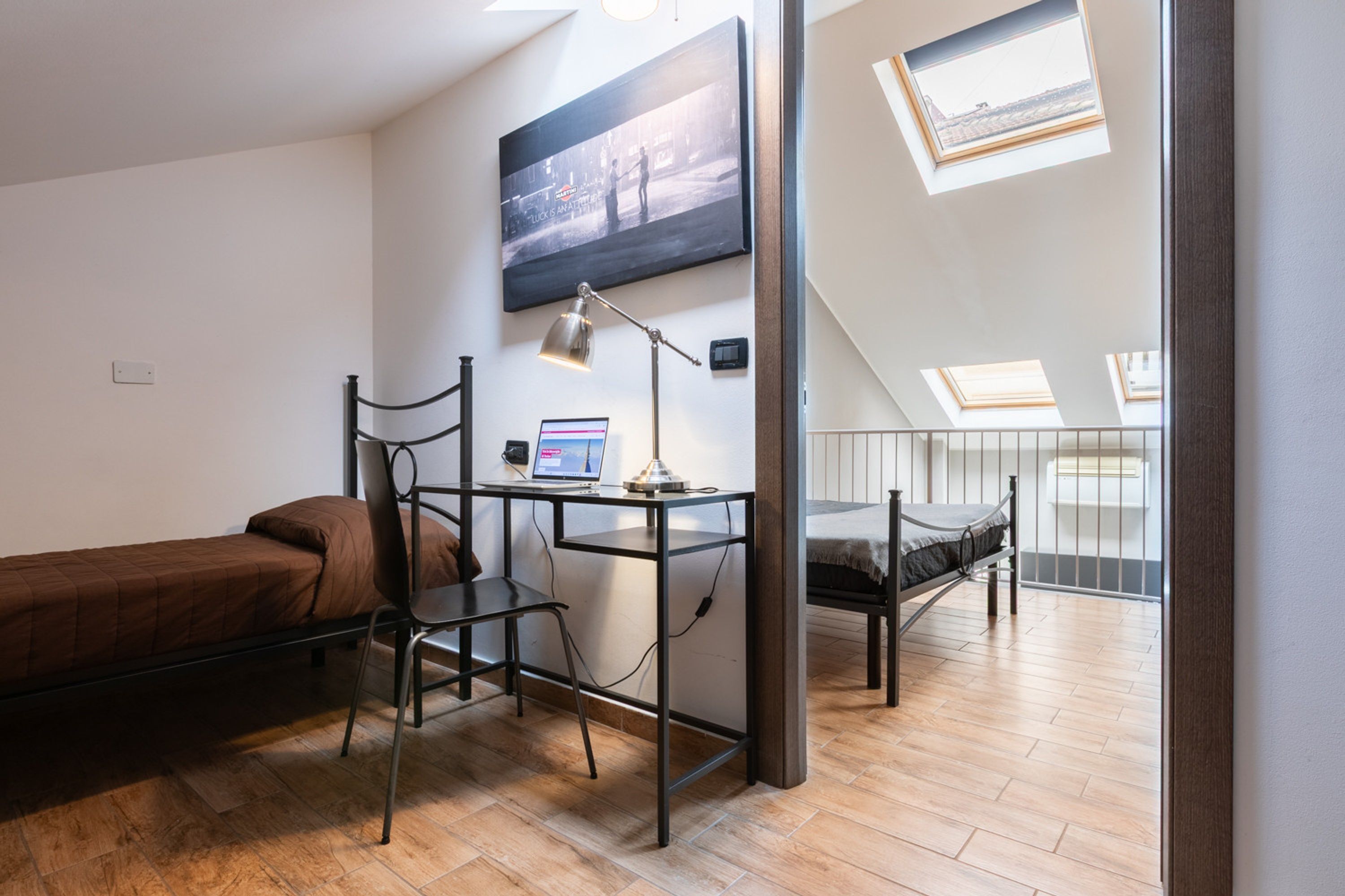 A compact room with a bed, desk, chair, and a laptop. There are skylights and a window providing natural light.
