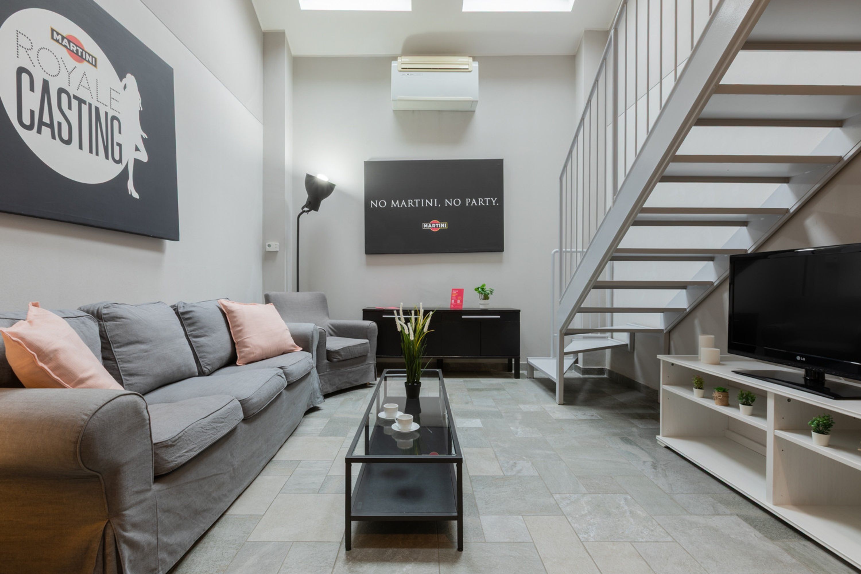 Un salon moderne avec un canapé gris, une table basse en verre, un tél éviseur à écran plat, un escalier et un mur orné d’une affiche de « Martini Royale Casting ».