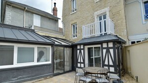 Eine überdachte Terrasse mit Tisch und Stühlen, ein Wintergarten mit Glastüren und ein Steinhaus mit Balkon.