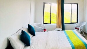 A hotel room with a bed, two white wicker chairs, a small table, and a window with curtains.
