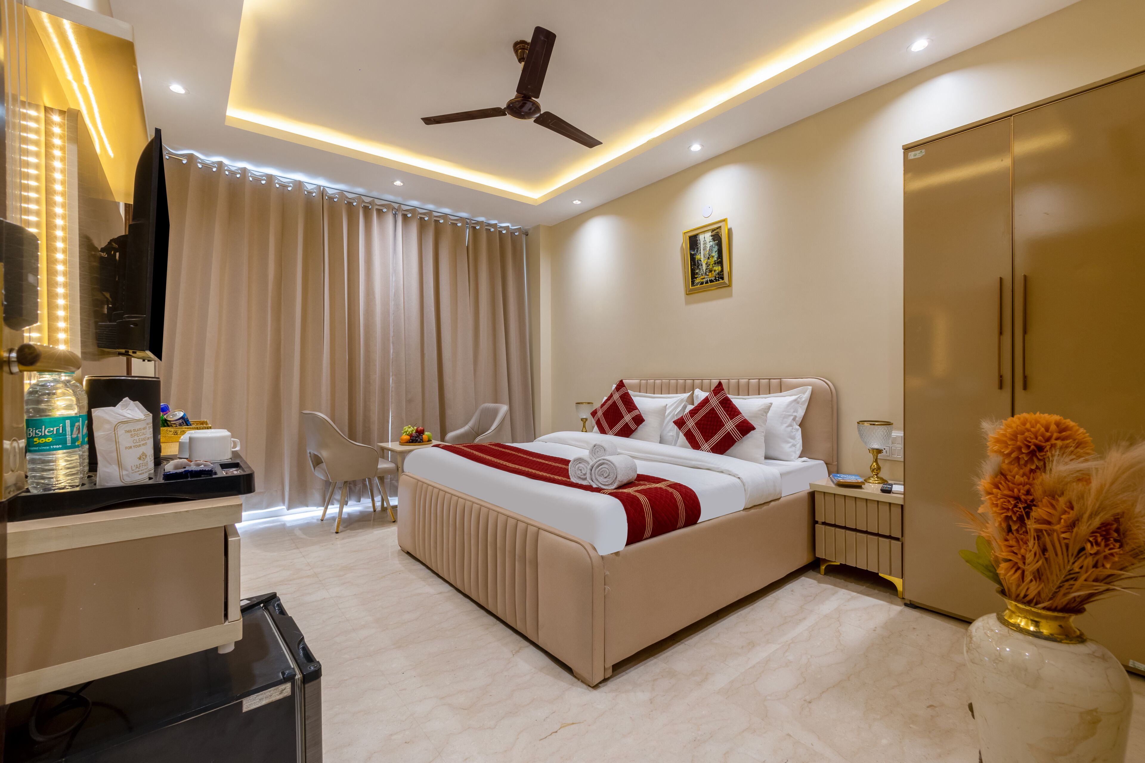 A hotel room with a bed, a desk, a chair, a wardrobe, a ceiling fan, and a vase with flowers.