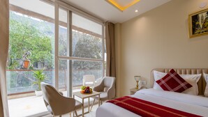 A hotel room with a large window, a bed with a red and white bedspread, a small table with fruit, and a chair. - Hotel Laffaire Select Near Rajori Metro (New Delhi)
