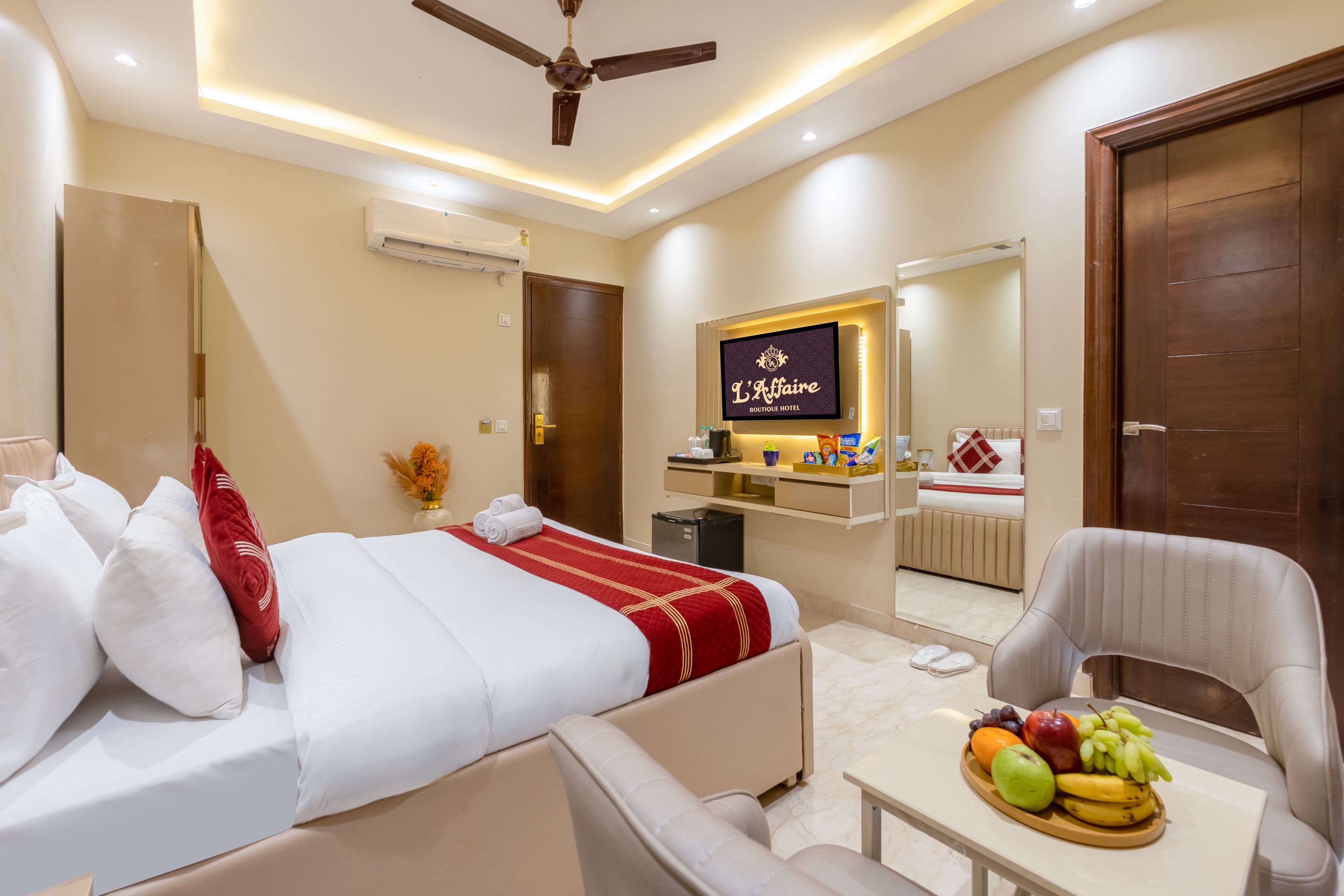 A hotel room with a bed, a desk with fruit, a chair, a ceiling fan, and a TV displaying 'L'Affaire Hotel'.