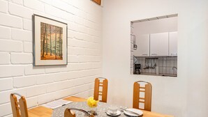 Een eetgedeelte met een houten tafel, vier stoelen, een lijst met een foto aan de muur, en een keuken zichtbaar door een raam.
