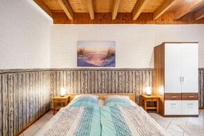 Una camera da letto con soffitto in legno, parete in mattoni bianchi, un quadro raffigurante un tramonto, un letto con lenzuola decorate, due comodini con lampade e un armadio.