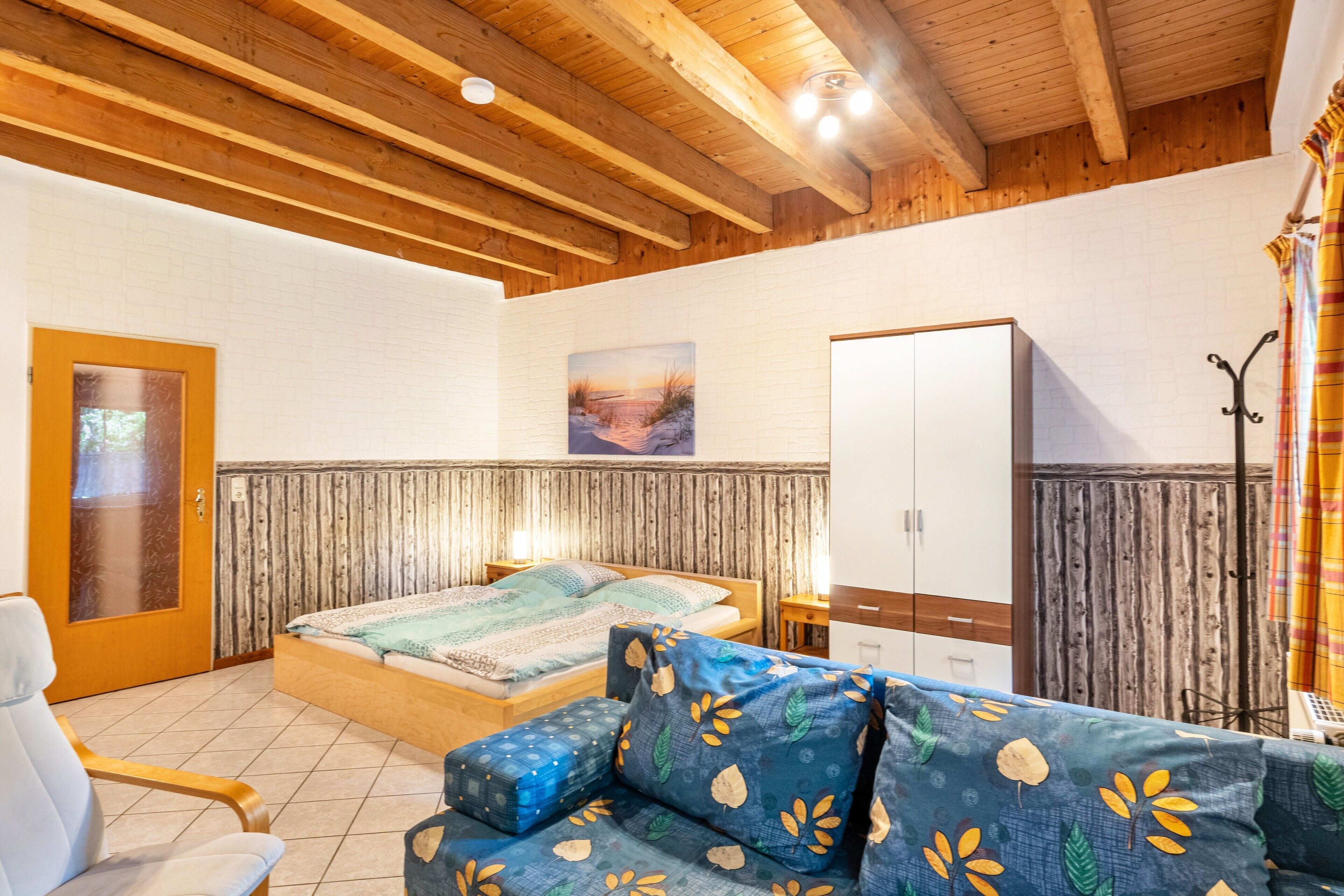 A room with a wooden ceiling, a bed with patterned bedding, a sofa with leaf designs, a wardrobe, and a painting on the wall.