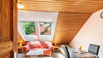 Ein gemütliches Dachzimmer mit schrägen Wänden, einem roten, gemusterten Sofa, einem kleinen Tisch und einem Esstisch mit floralem Tischläufer.