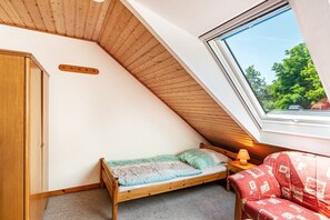 A small, sloped-roofed room with a single bed, a wooden wardrobe, a red sofa, and a window offering a view of greenery. - Vacation at the uhlenhoff apartment hieve (Ihlow)