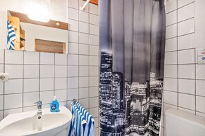 Un baño con un lavamanos blanco, una botella azul de jabón para manos, una toalla a rayas y una cortina de ducha con diseño de paisaje urbano.