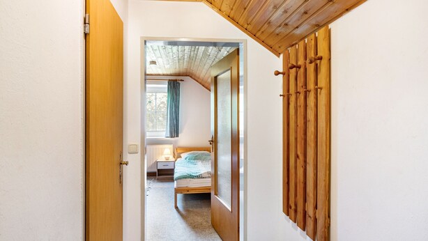 A narrow hallway leading to a bedroom with a wooden ceiling, a bed, and a window with curtains.