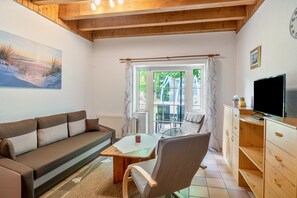 Ein gemütliches Wohnzimmer mit einer Couch, einem Holztisch, einem Sessel, einem Fernseher und einem Fenster mit Vorhängen.