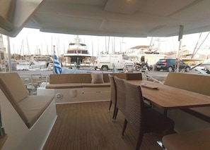Un espace extérieur abrité, aménagé avec des tables et des chaises, une terrasse en bois et une marina où l’on aperçoit des bateaux au loin.