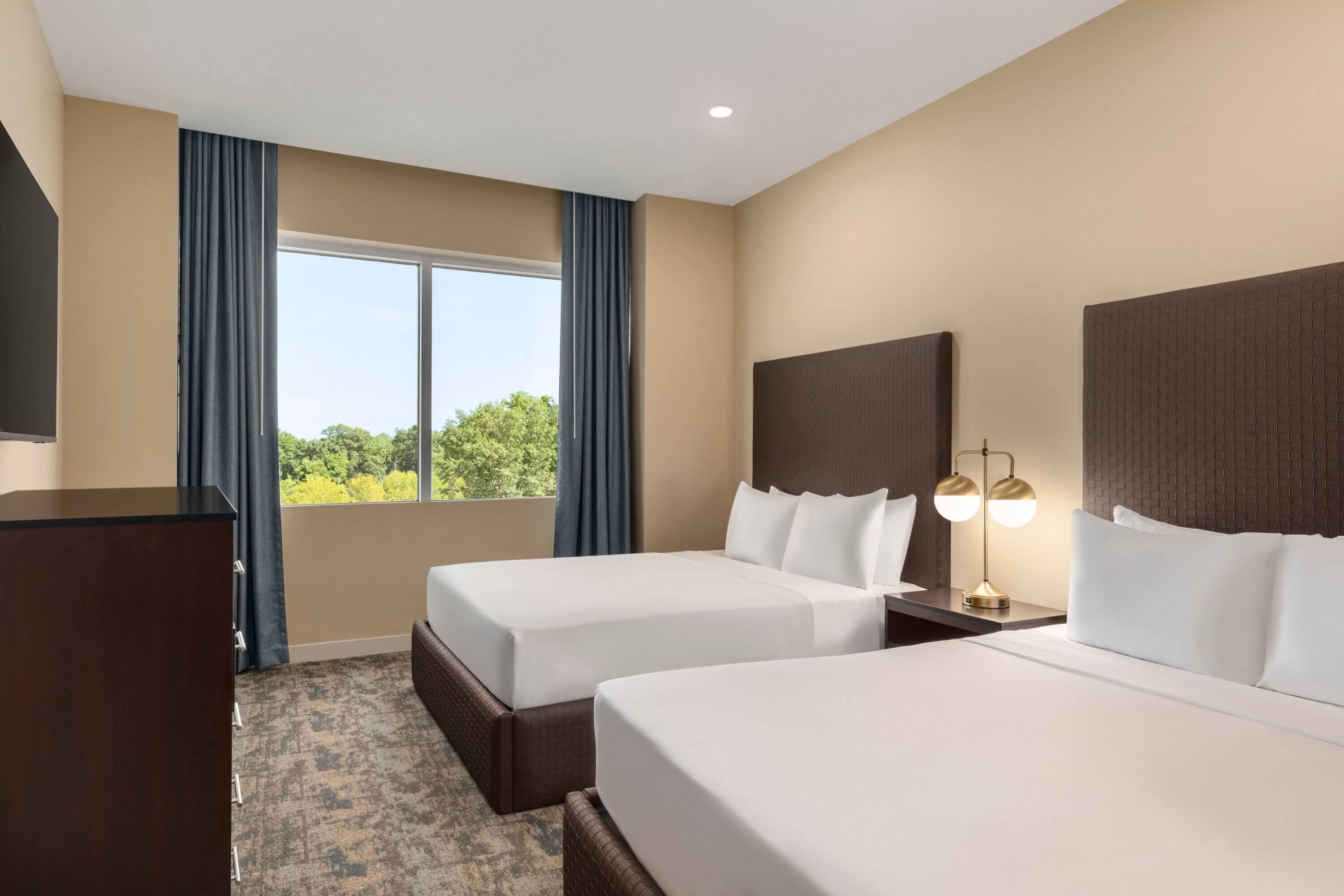A hotel room with two beds, a desk, a lamp, and a view of trees through the window.