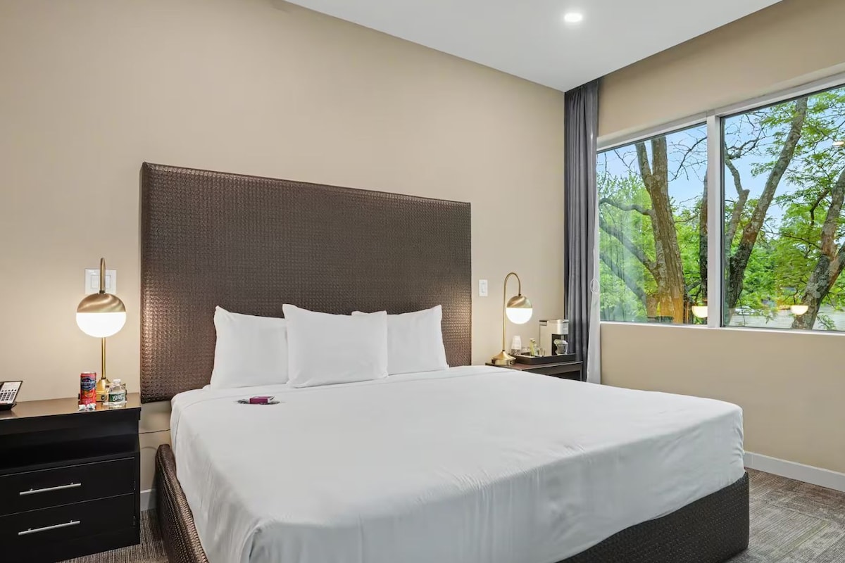 A hotel room with a large bed, a nightstand, a desk, and a window with a view of trees.
