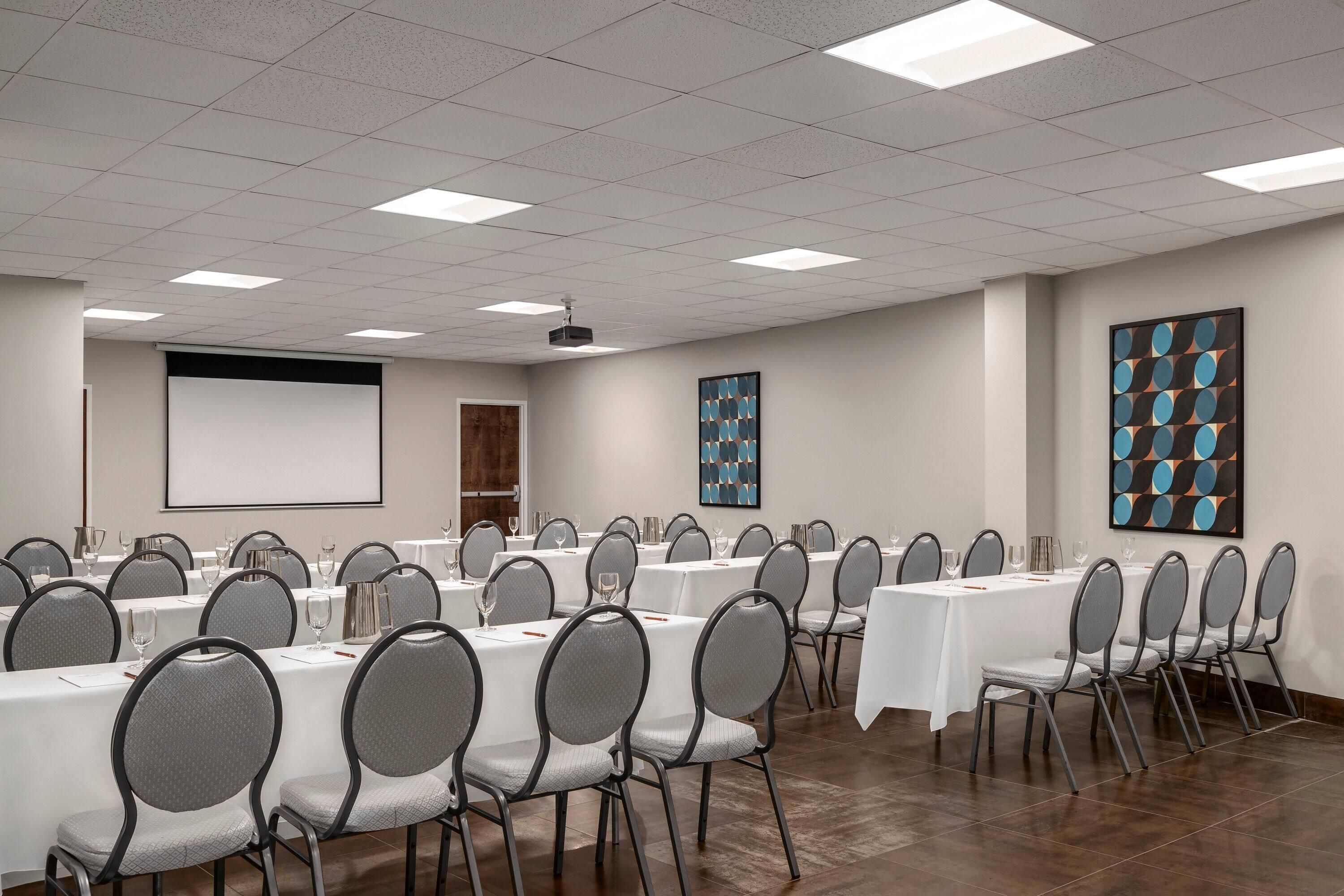 A conference room with multiple tables and chairs, a projector screen, and abstract art on the walls.
