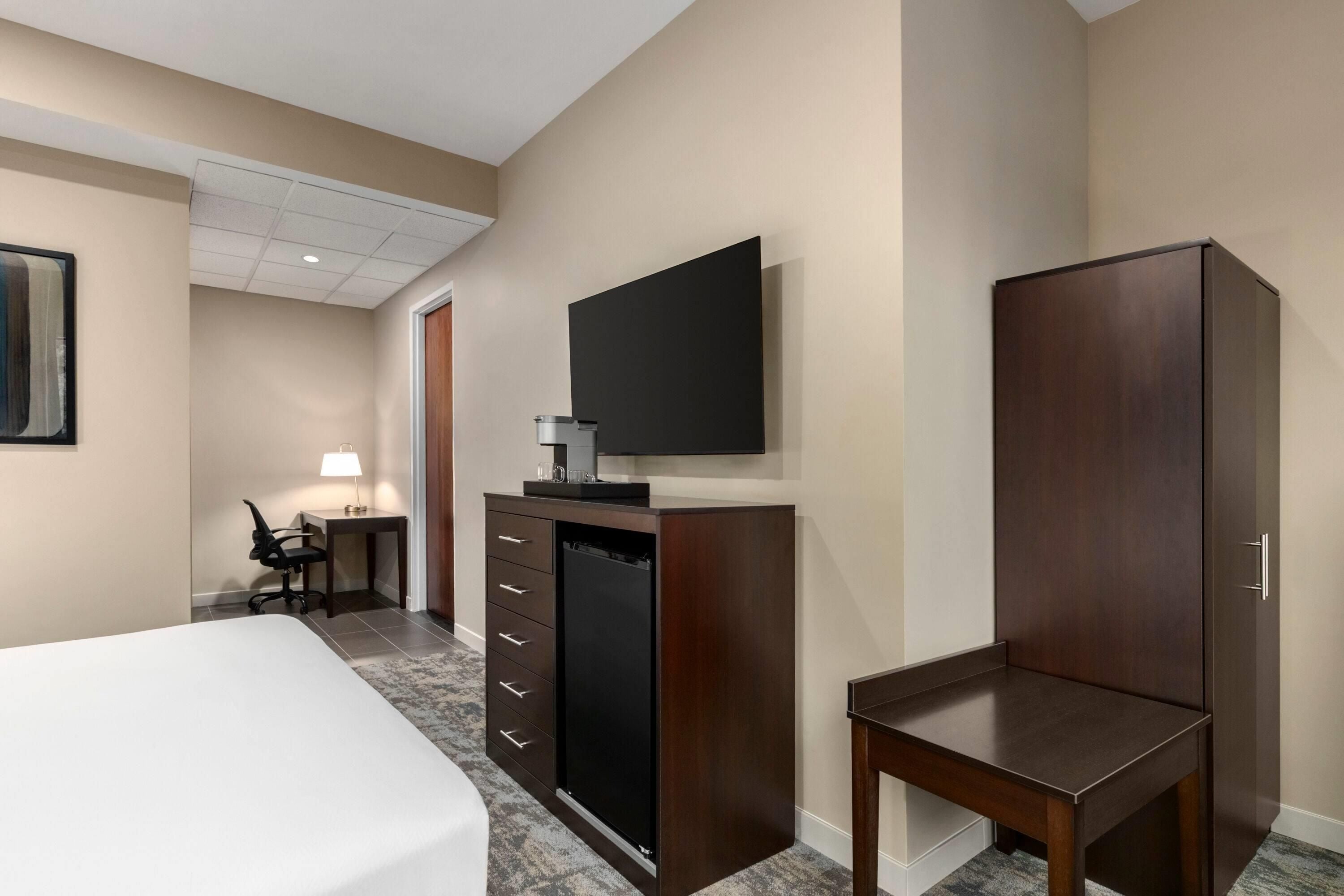 A hotel room with a bed, a desk with a lamp, a TV mounted on the wall, and a wardrobe.