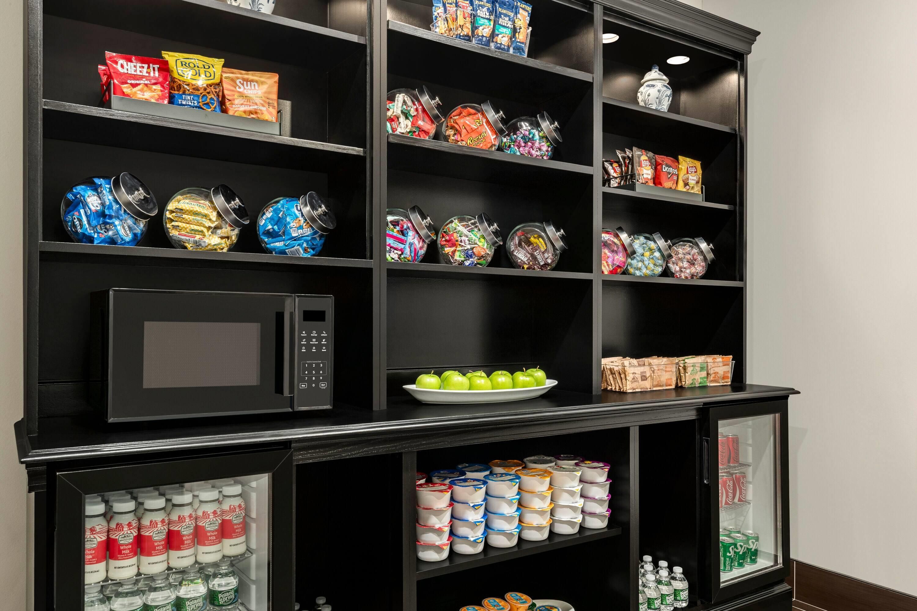 A kitchenette with a microwave, shelves stocked with snacks, a plate of green apples, and a refrigerator containing beverages and dairy products.
