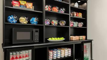 A kitchenette with a microwave, shelves stocked with snacks, a plate of green apples, and a refrigerator containing beverages and dairy products.