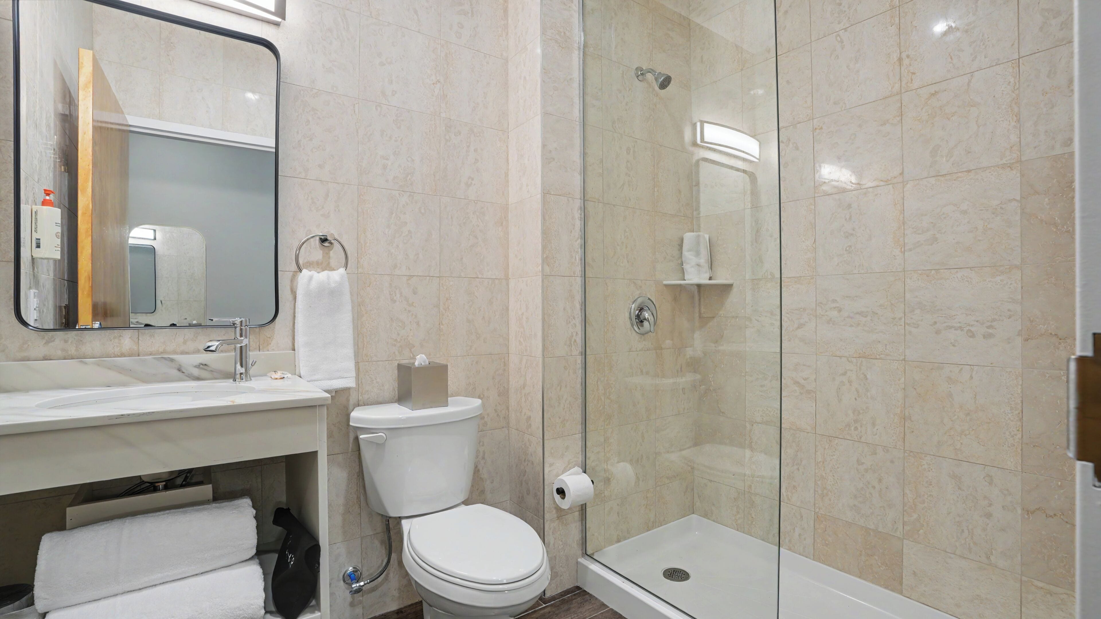 Un baño con inodoro, lavamanos, espejo, toallero y cabina de ducha con mampara de vidrio.