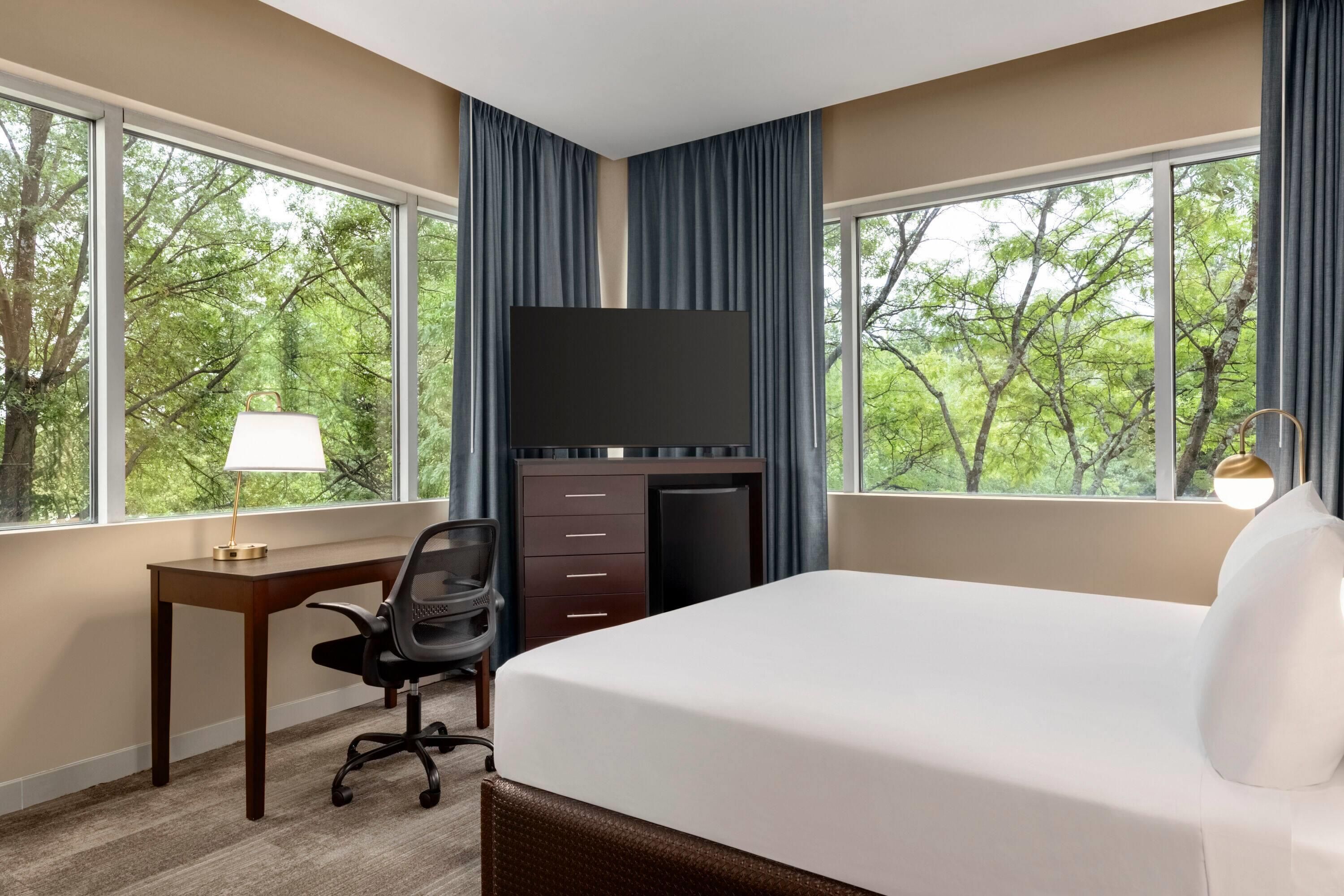 A hotel room with a bed, desk, chair, TV, and large windows with curtains.