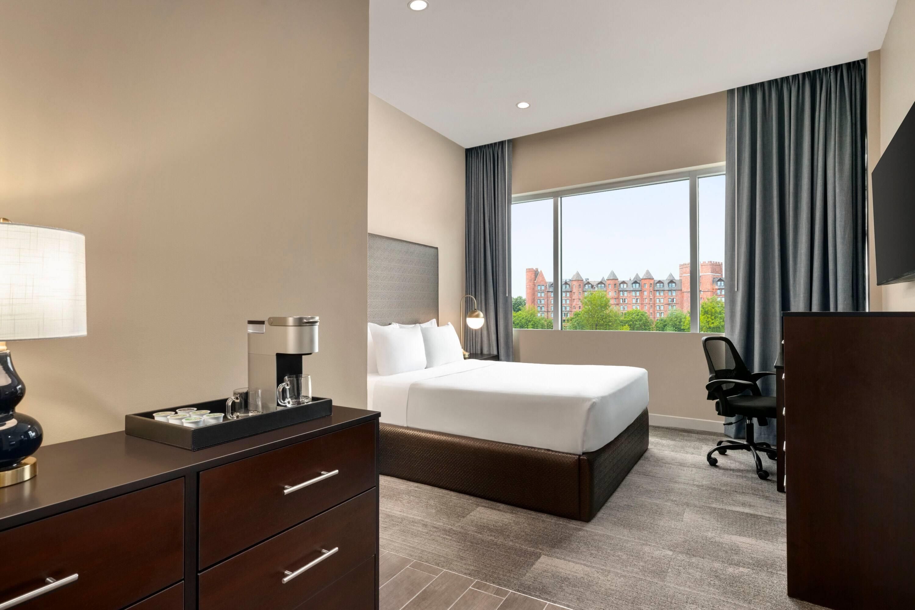 A hotel room with a bed, a desk with a coffee maker, a chair, and a large window with curtains.