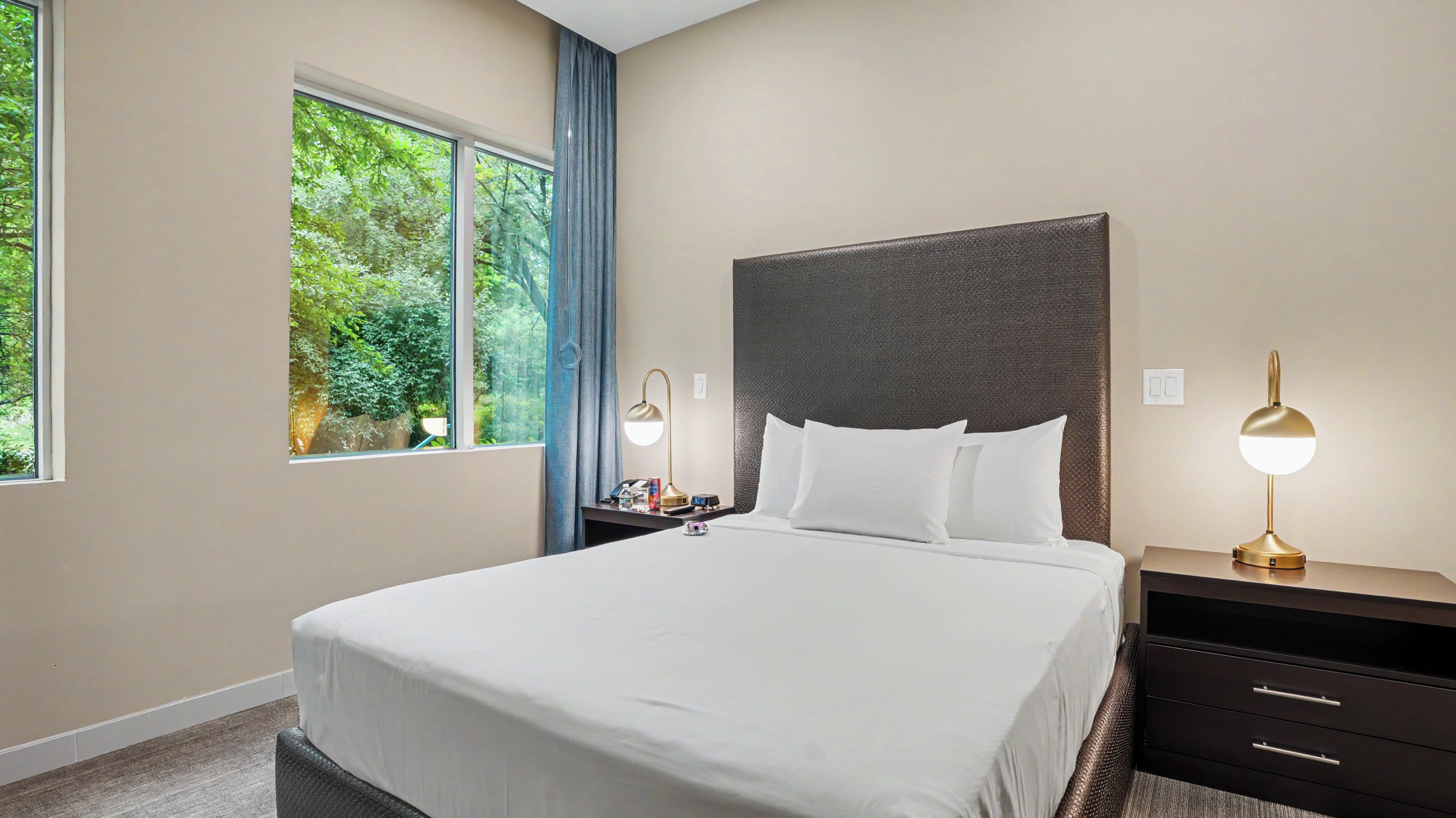 A hotel room with a large bed, two bedside tables, a nightstand, a window with a view of greenery, and a wall-mounted lamp.