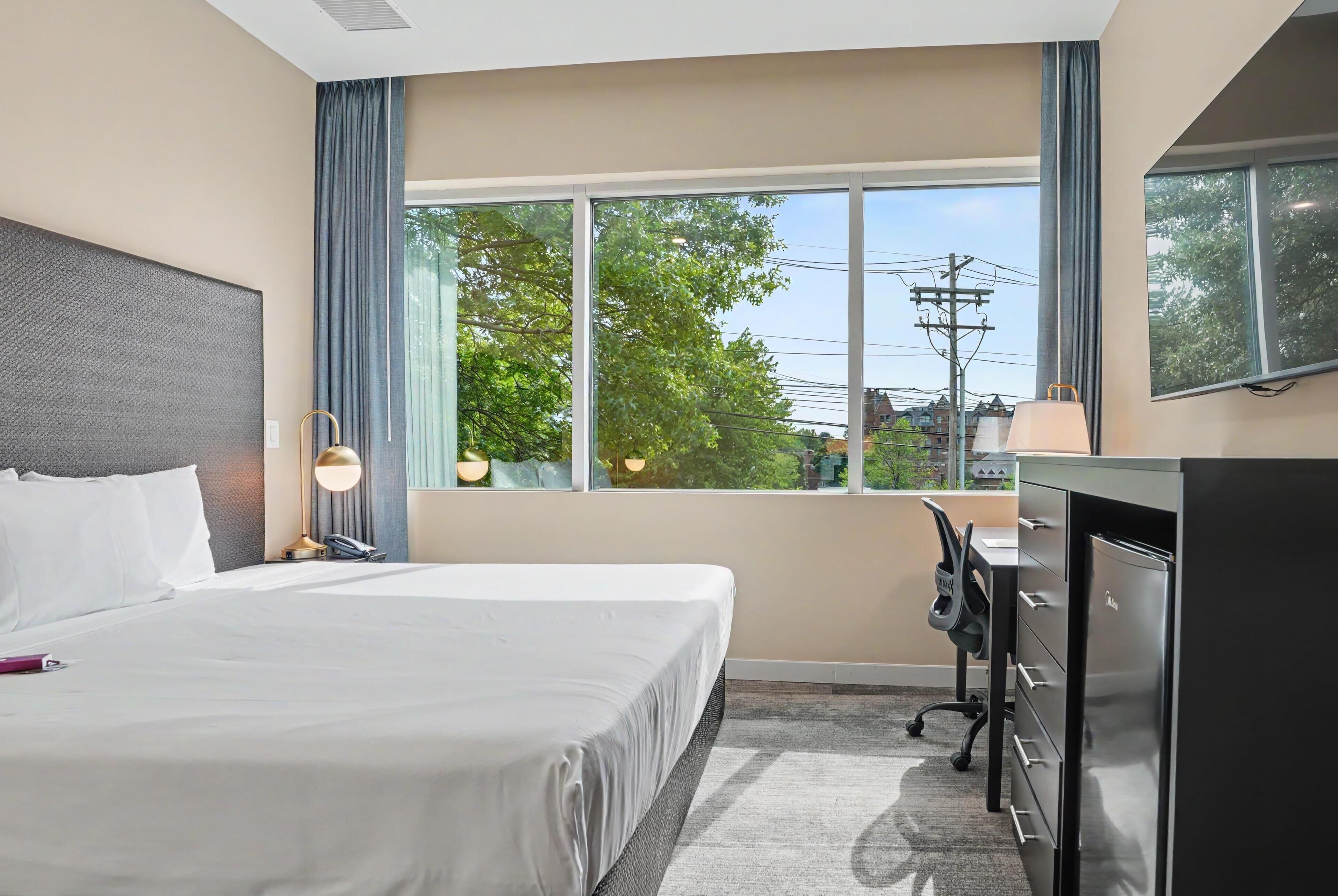 A hotel room with a bed, desk, chair, and a view of trees and buildings.