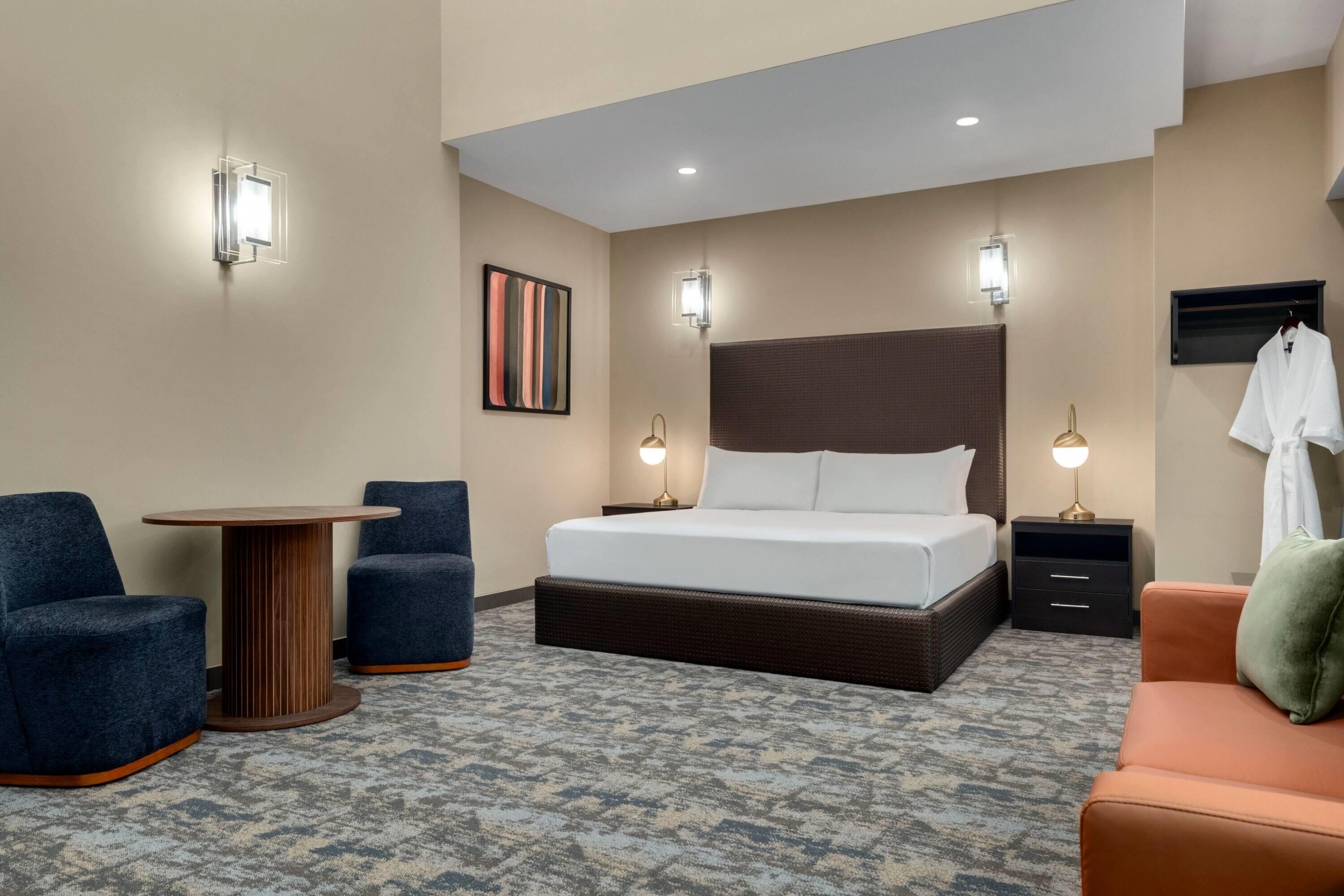 A hotel room with a bed, two chairs, a small table, a TV, and a robe hanging on the wall.