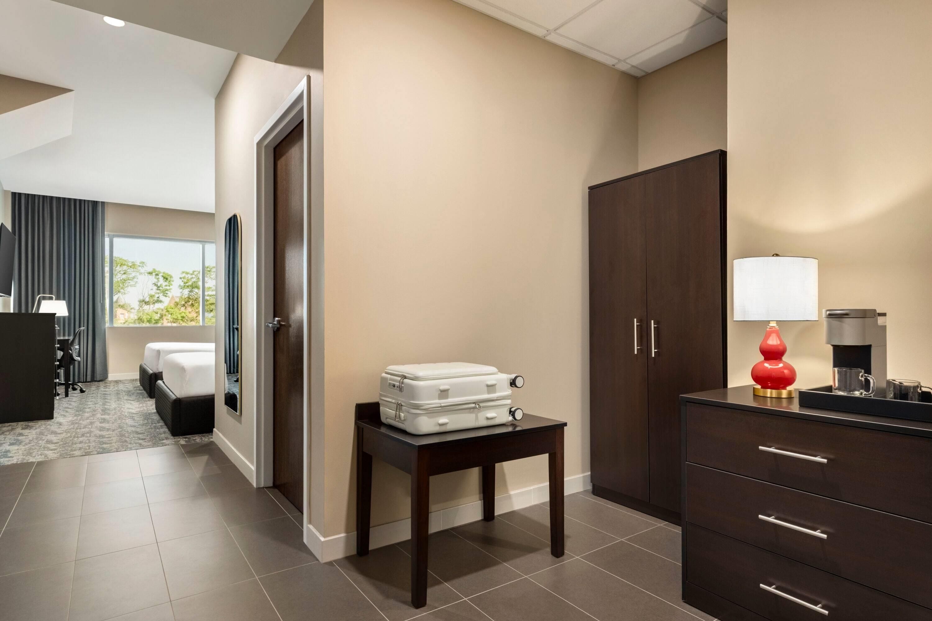 A hotel room with a bed, a desk, a wardrobe, and a table with a suitcase.