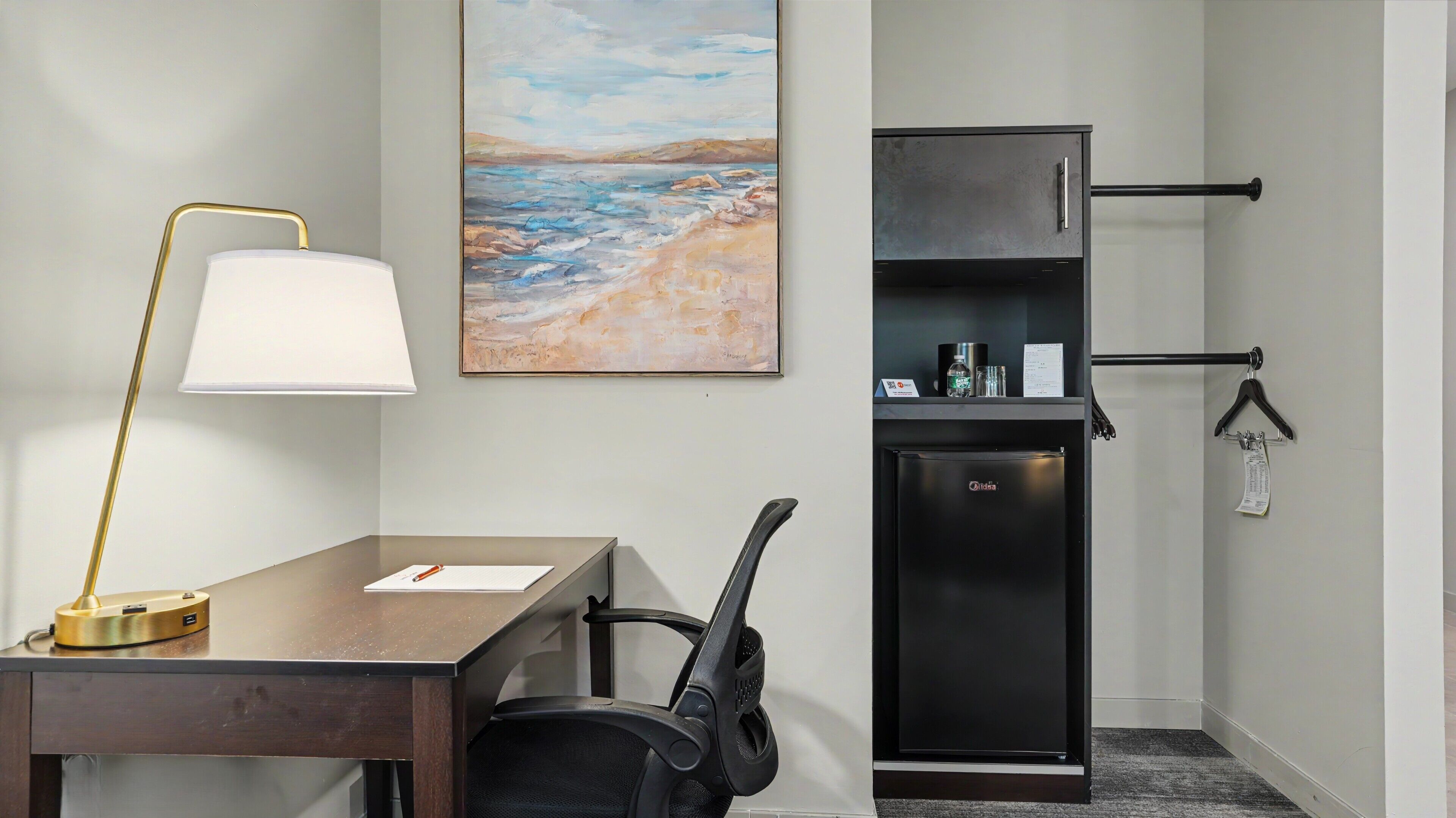 Habitación de hotel con escritorio, silla, lámpara, refrigerador y una obra de arte en la pared.