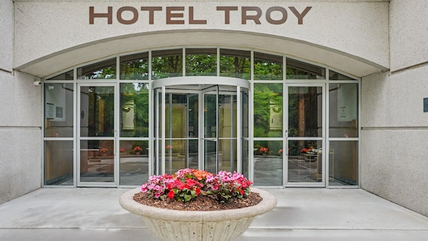 L’entrée d’un hôtel, avec une porte tournante, une enseigne indiquant « HÔTEL TROY » et un parterre de fleurs devant.