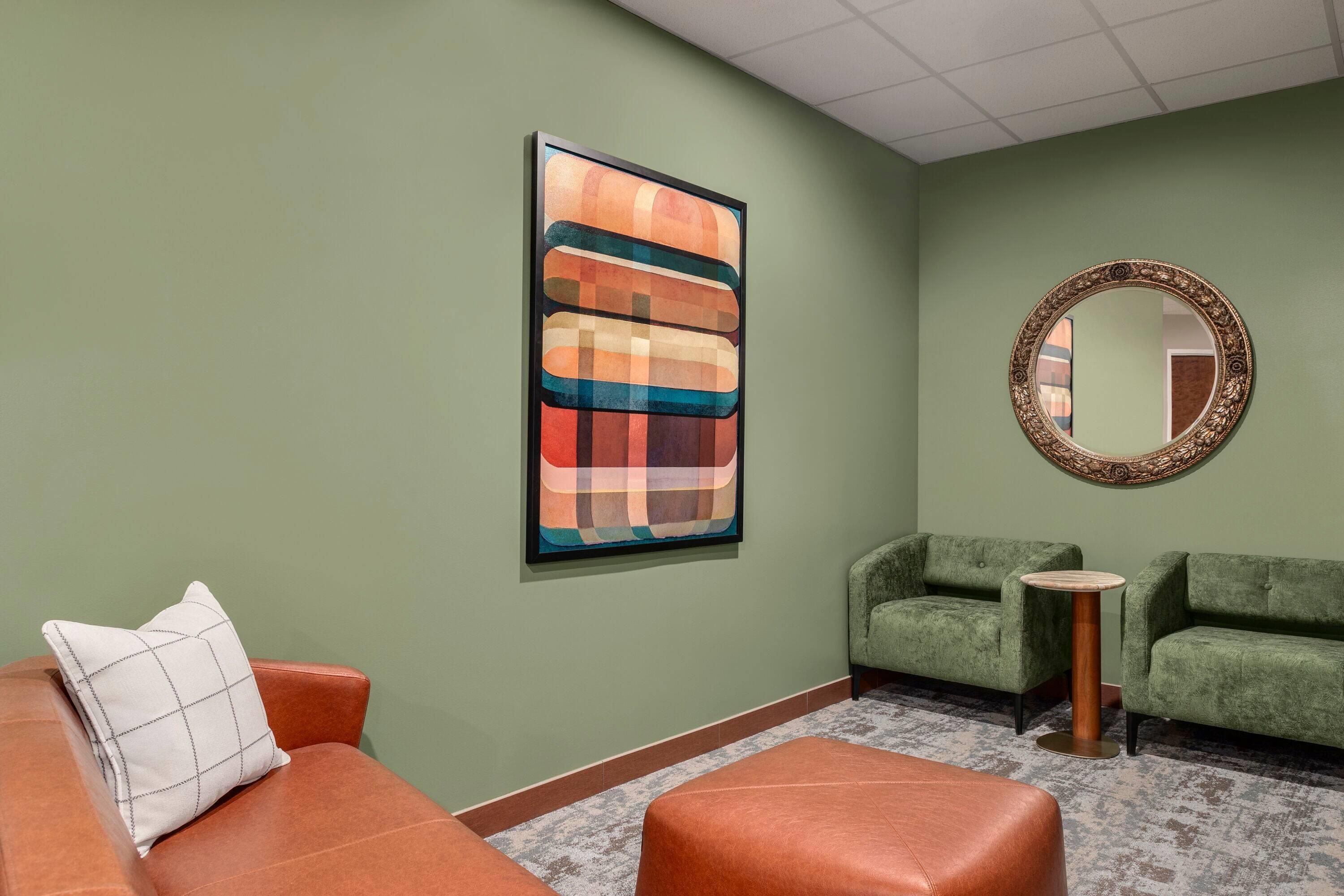 Una habitación con pared verde, un cuadro abstracto enmarcado, un espejo redondo, dos sofás verdes, una mesita redonda y un otoman de cuero marrón.