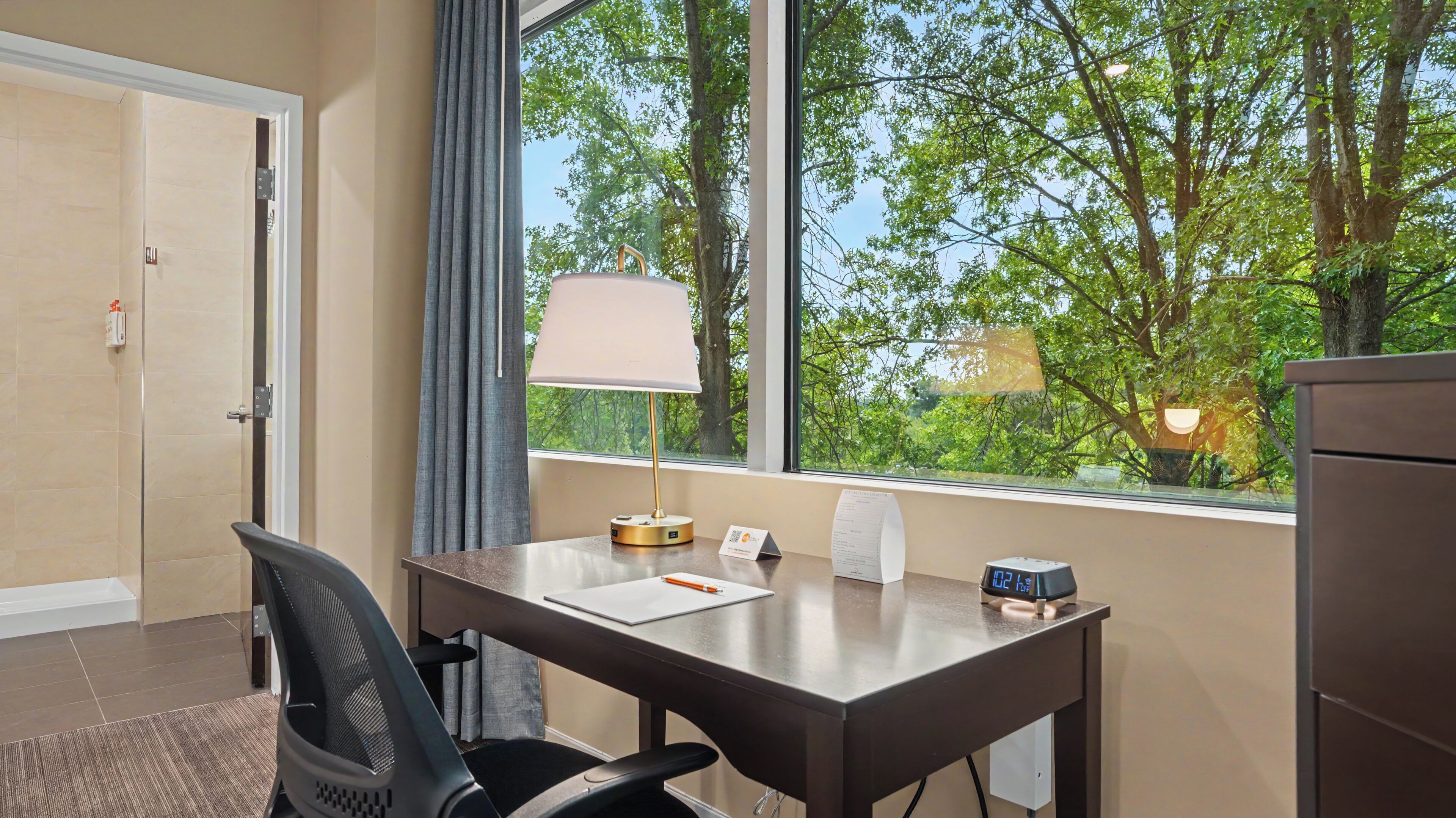 A hotel room with a desk, chair, lamp, clock, and a window with a view of trees.
