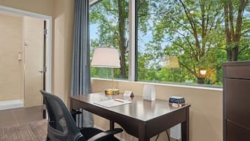 A hotel room with a desk, chair, lamp, clock, and a window with a view of trees.