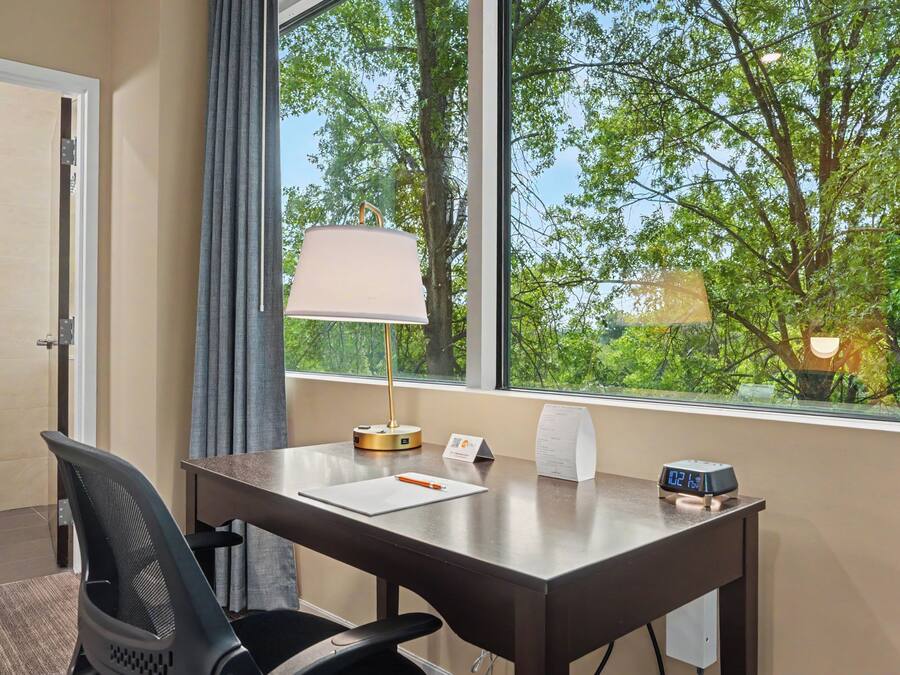 A hotel room with a desk, chair, lamp, clock, and a window with a view of trees.