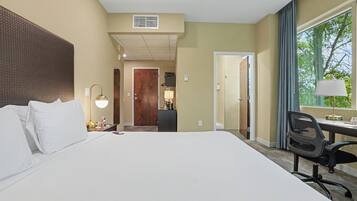 Habitación de hotel con una cama grande, un escritorio con silla, una ventana con vista a árboles y una puerta que da a un baño.