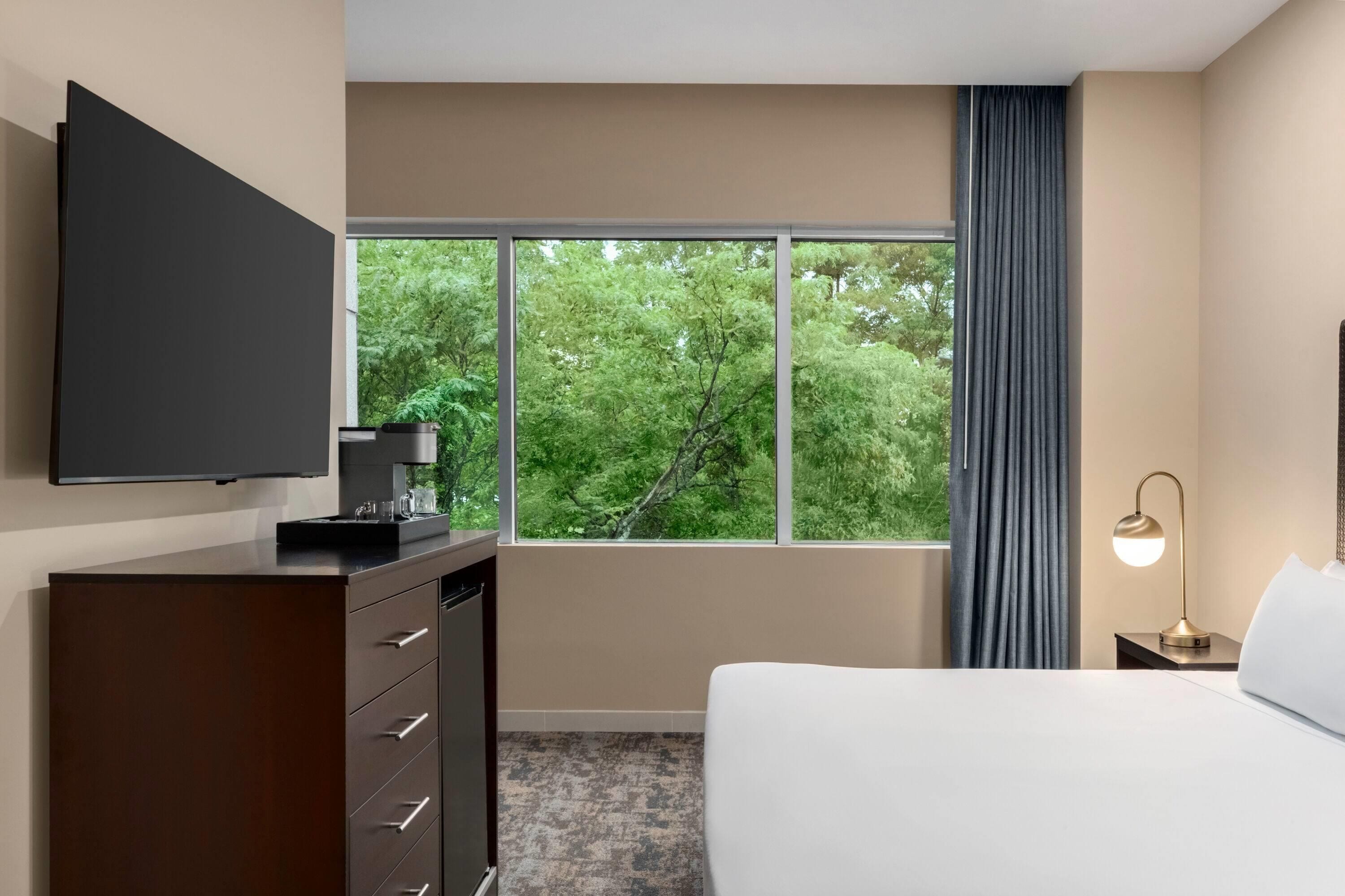 Une chambre d’hôtel avec un lit, un téléviseur fixé au mur, une machine à café, une lampe et une vue sur les arbres par la fenêtre.