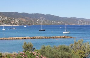A coastal town with boats on the water, rocky breakwater, and a hillside with buildings.