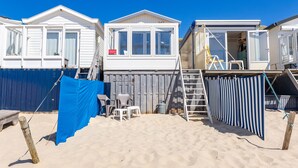 Una casa móvil frente a la playa con un porche delantero, un pequeño espacio de estar al aire libre y una escalera que lleva al nivel superior.